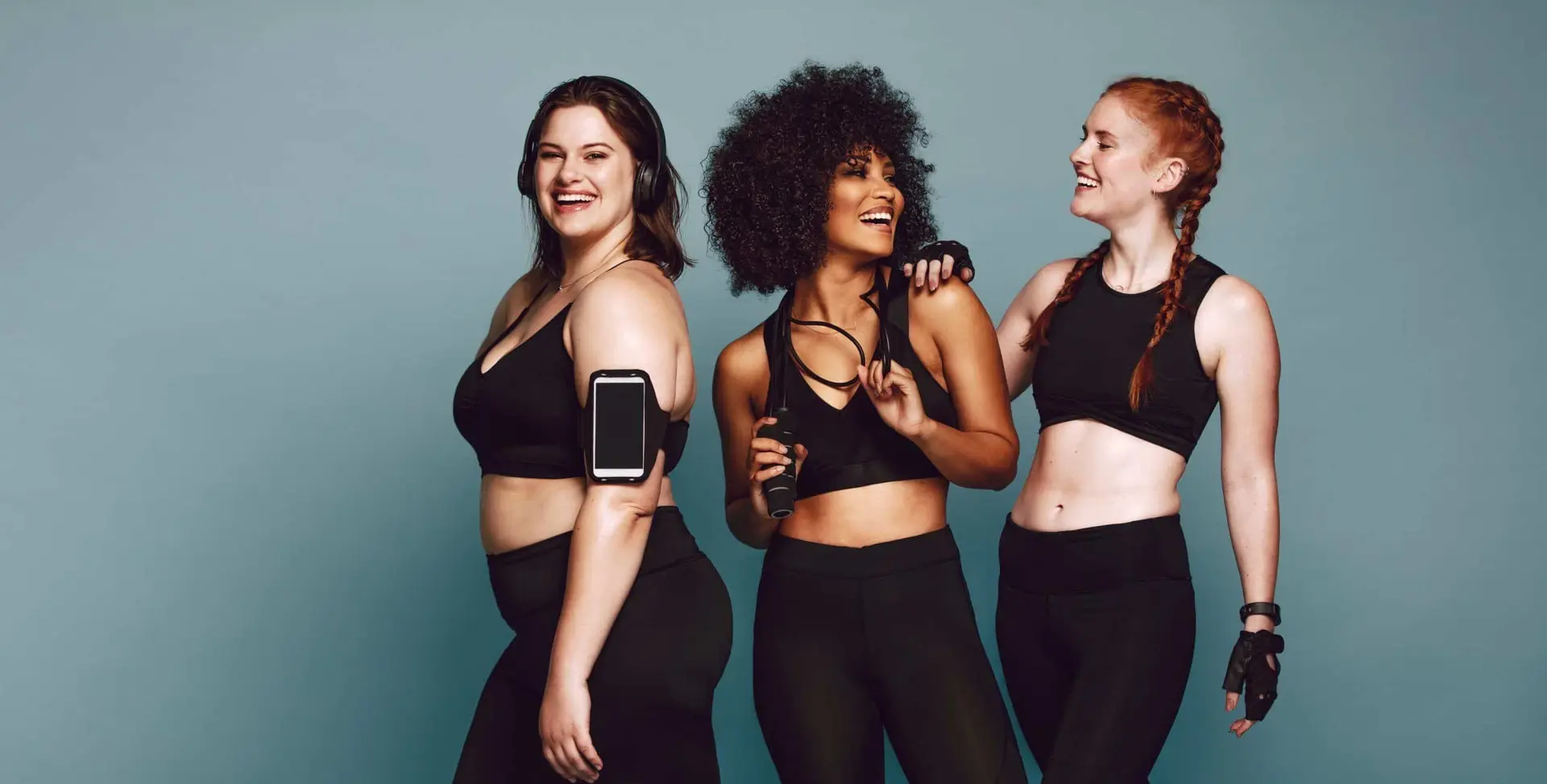 Drie vrouwen in zwarte sportkleding lachen samen; een draagt een koptelefoon en de smartphone om de bovenarm, een houdt een springtouw vast